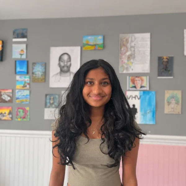Our Team 68 A smiling young woman with long, voluminous black hair wearing an olive-gray sleeveless top and delicate gold jewelry, standing in front of a gray gallery wall decorated with colorful artwork, portraits, and sketches