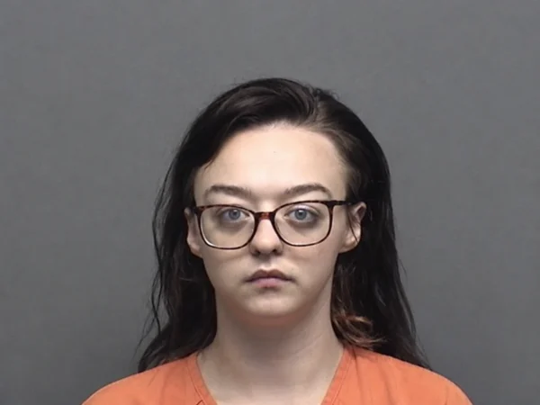 Kiera, A young woman with long dark hair and rectangular glasses looks directly at the camera with a neutral expression, wearing an orange shirt against a plain gray background.