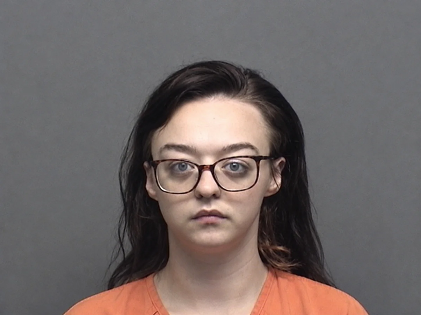 Kiera, A young woman with long dark hair and rectangular glasses looks directly at the camera with a neutral expression, wearing an orange shirt against a plain gray background.