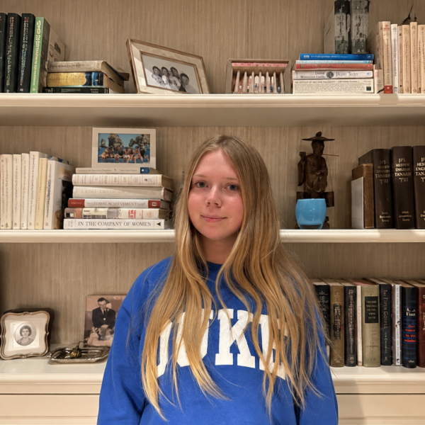 Our Team 67 A young woman with long, straight blonde hair and a gentle smile wears a royal-blue Duke sweatshirt while standing in front of cream-colored bookshelves filled with books, framed photographs, a blue vase, and decorative sculptures.