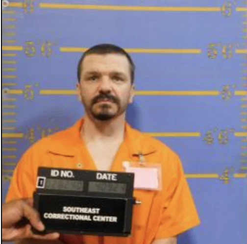 Mugshot of a man with short dark hair, a mustache, and a goatee, wearing an orange jumpsuit in front of a blue height chart backdrop labeled "Southeast Correctional Center.