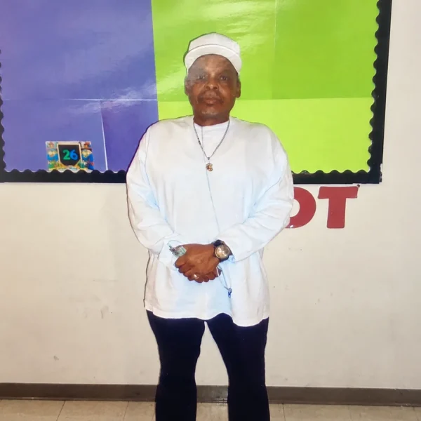 Our Team 36 A man wearing a white cap and traditional white tunic with dark trousers, a necklace, and a wristwatch, standing in front of a colorful classroom bulletin board.