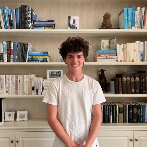 Our Team 71 Smiling young man with curly dark hair wearing a white T-shirt and silver chain, standing with his hands clasped in front of a cream-colored bookshelf filled with books, framed photographs, and decorative objects.