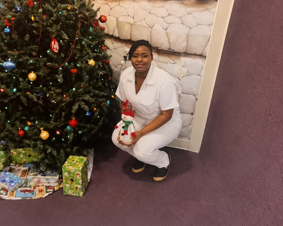 Amesty, 30 3 Woman in white outfit holding snowman plush beside decorated Christmas tree with gifts.